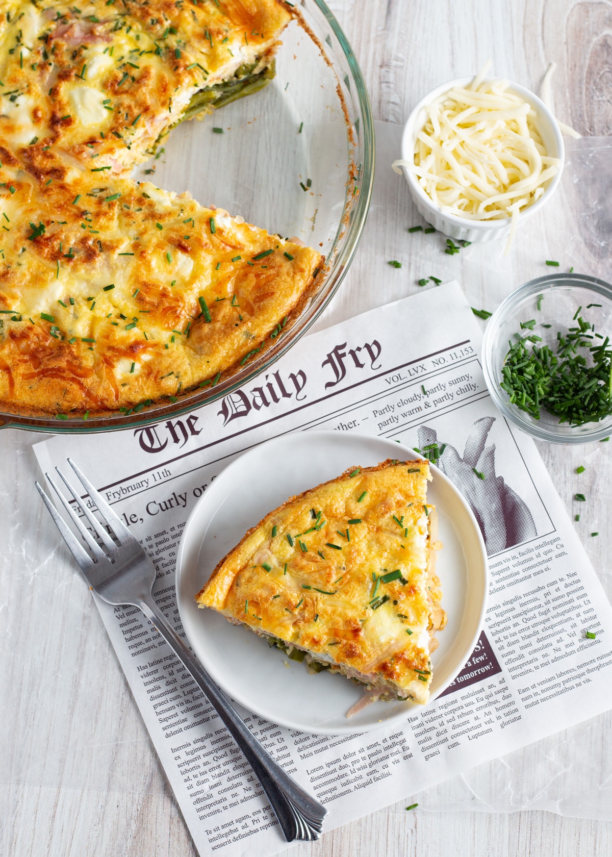 Picture with a slice of crustless quiche with asparagus ham and goat cheese with rest of quiche in background and little bowls with cheese and chives. 