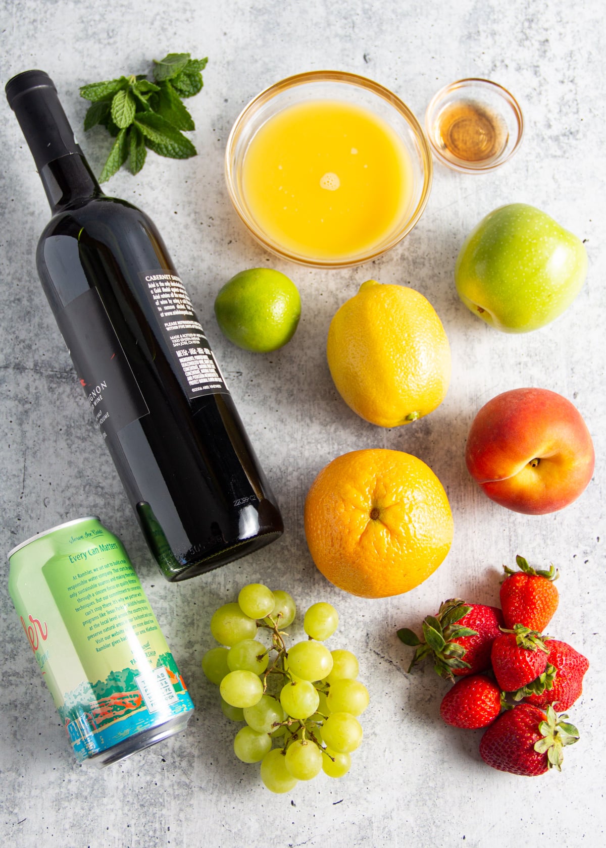 Picture of each individual ingredients for non-alcoholic sangria: alcohol-removed wine, orange juice, agave, orange, lemon, lime, strawberries, apple, grapes and lemon-lime seltzer.