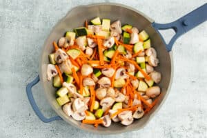 Picture of raw mushrooms, zucchini and carrots added to skillet to cook.