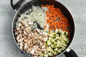 This is a picture of a skillet with onion, carrots, mushrooms and zucchini cooking.
